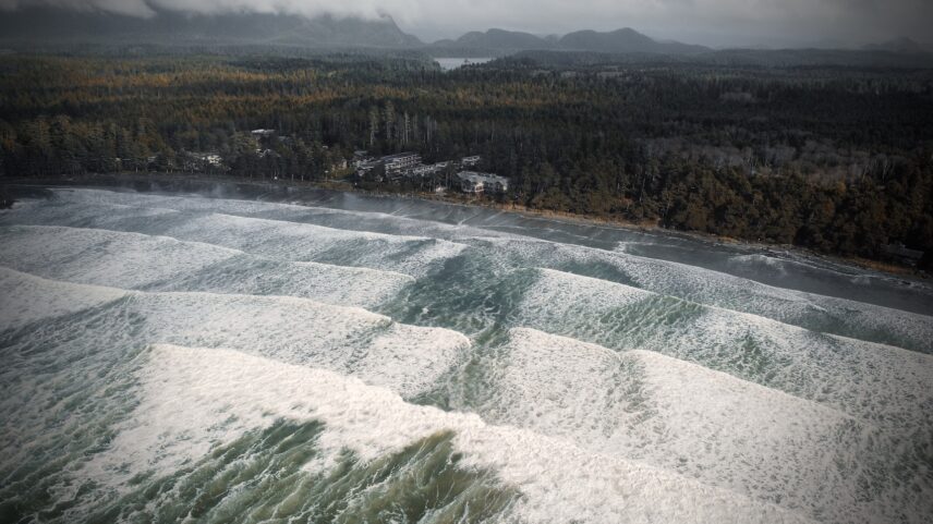 Dramatic aerial of Cox Bay in Tofino, showing powerful incoming waves and a stormy ocean.